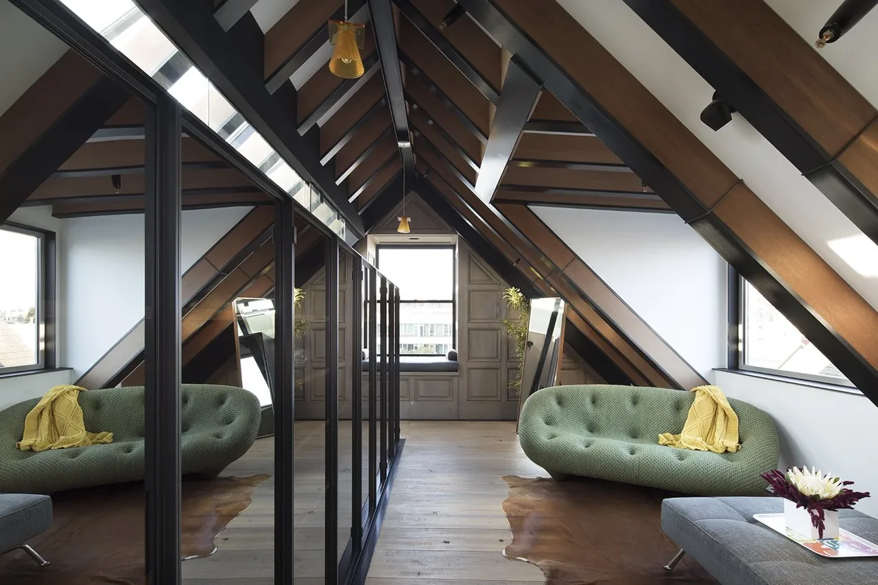 Living area under sloped ceiling with wooden beams and window bench, reflecting in glass partition.