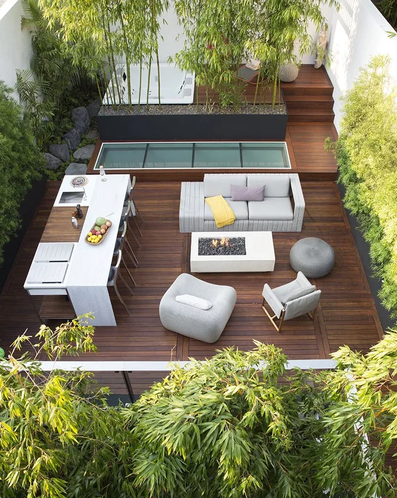 Modern outdoor living space features a wooden deck with seating, fire pit, and kitchen island surrounded by bamboo.