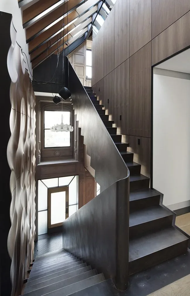 A staircase featuring a black steel railing and wood-paneled walls with sculptural wall detailing, illuminated by natural light from overhead skylights.