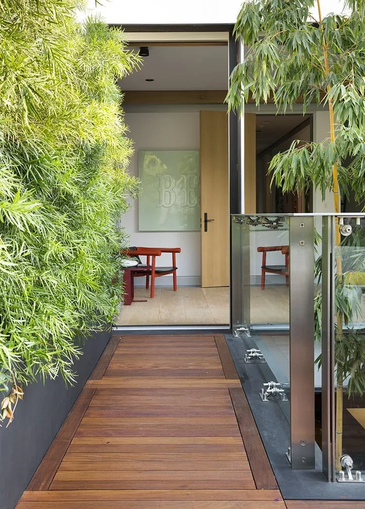 A wood plank deck walkway bordered by a planter and glass railing leads to an entrance with minimalist modern furniture inside.