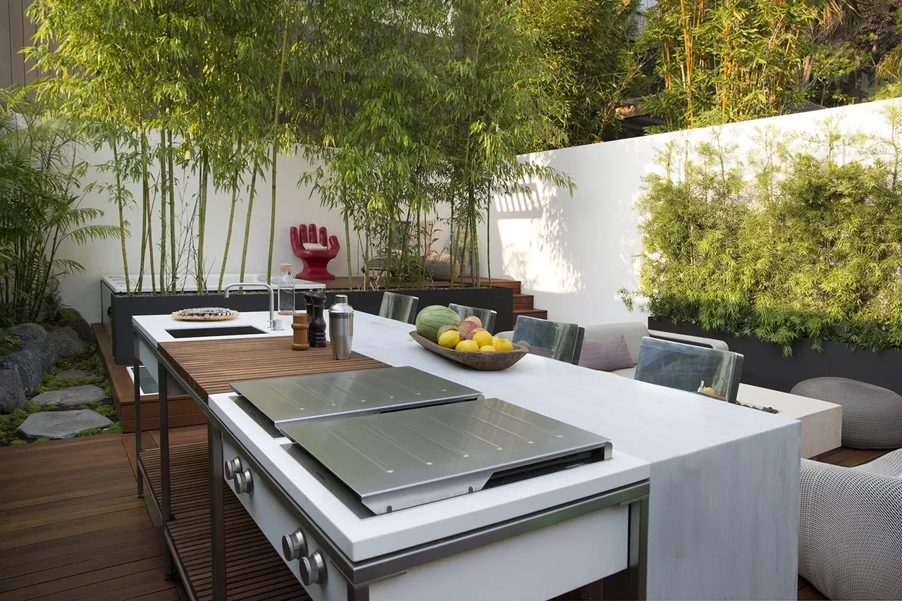 Modern outdoor kitchen and seating area in a backyard with lush plants and wooden deck flooring.
