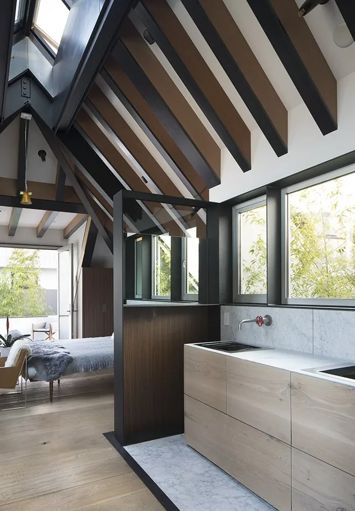 Open kitchen with light wood cabinets under a sloped, beamed ceiling and large windows revealing greenery.