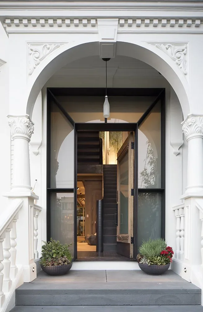 Exterior front view of a white building with a large black-framed arched glass door revealing an interior staircase.