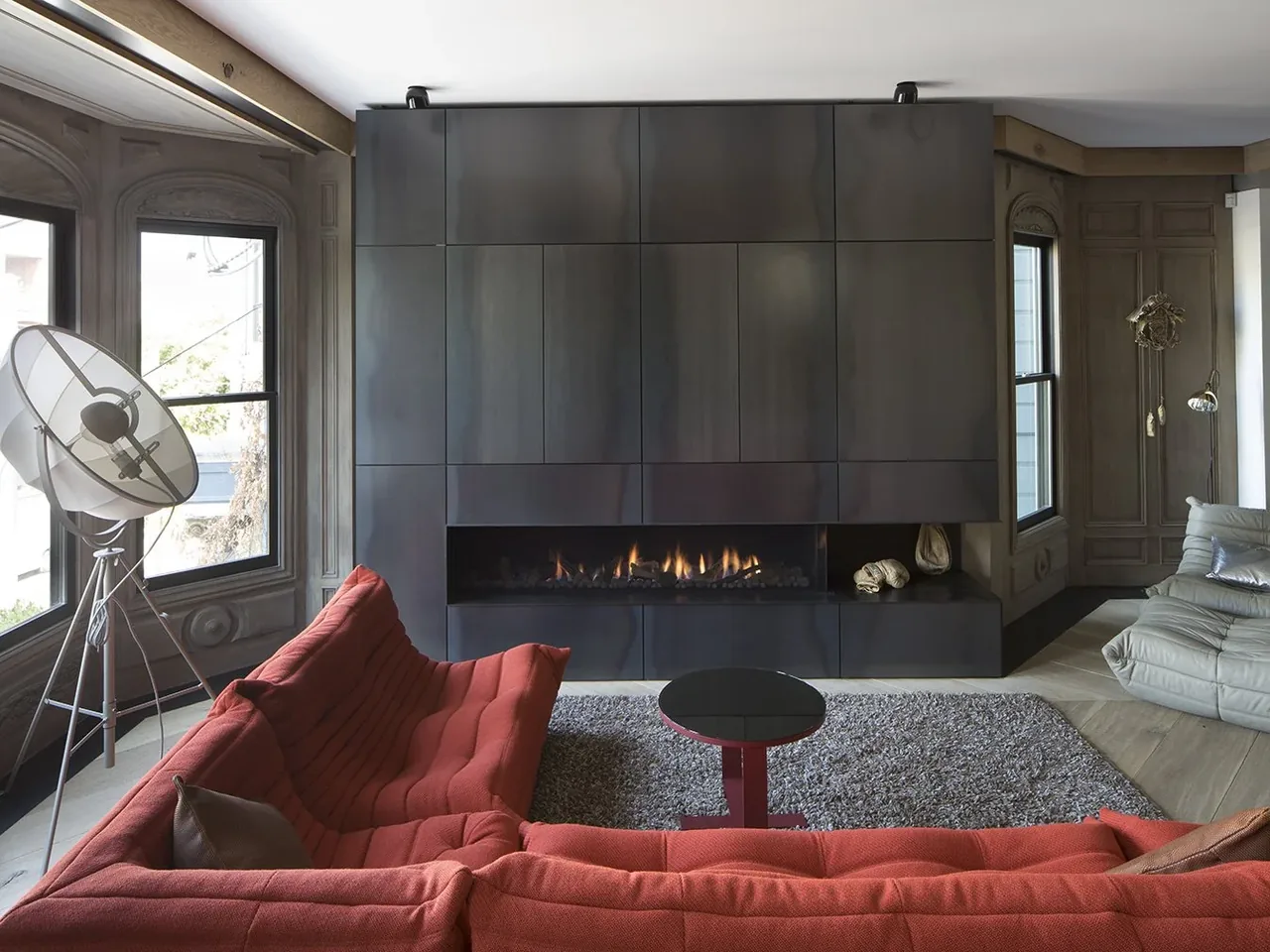 Living room featuring a black metal-paneled fireplace wall with a gas fire, red sectional sofa, and a large tripod floor lamp near windows.