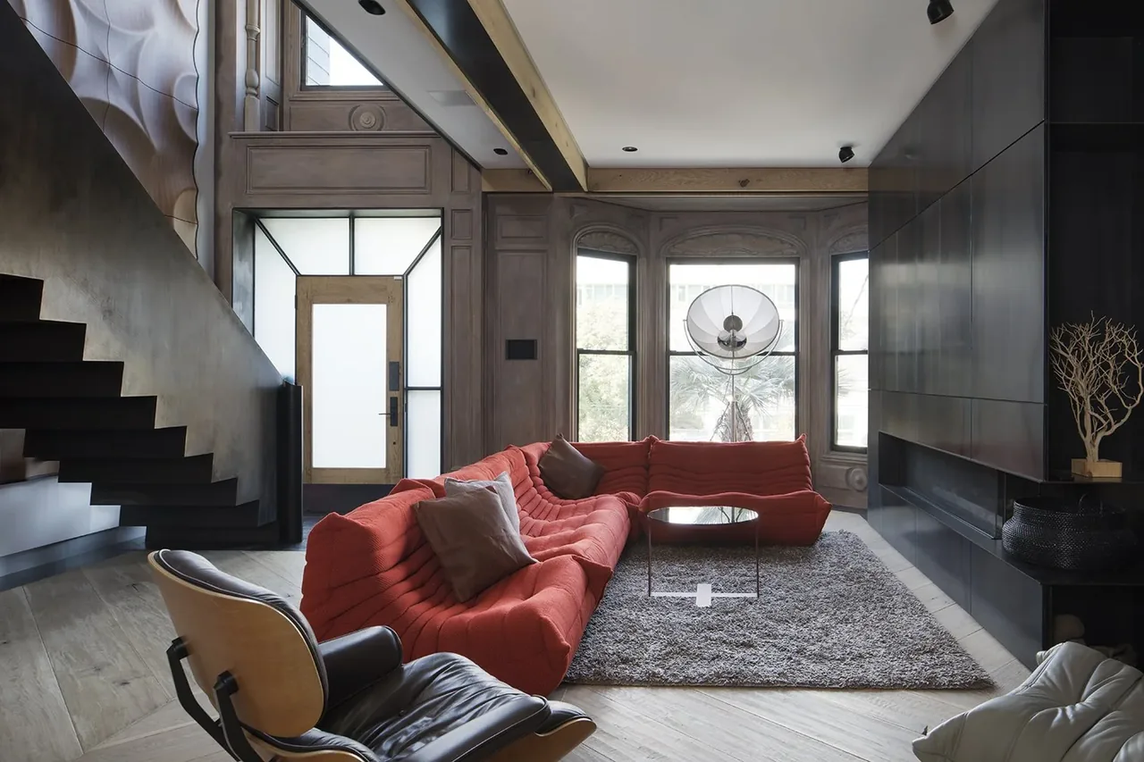 Living room with a red sectional sofa, textured wall panel, and black cabinets next to large windows and a modern staircase.