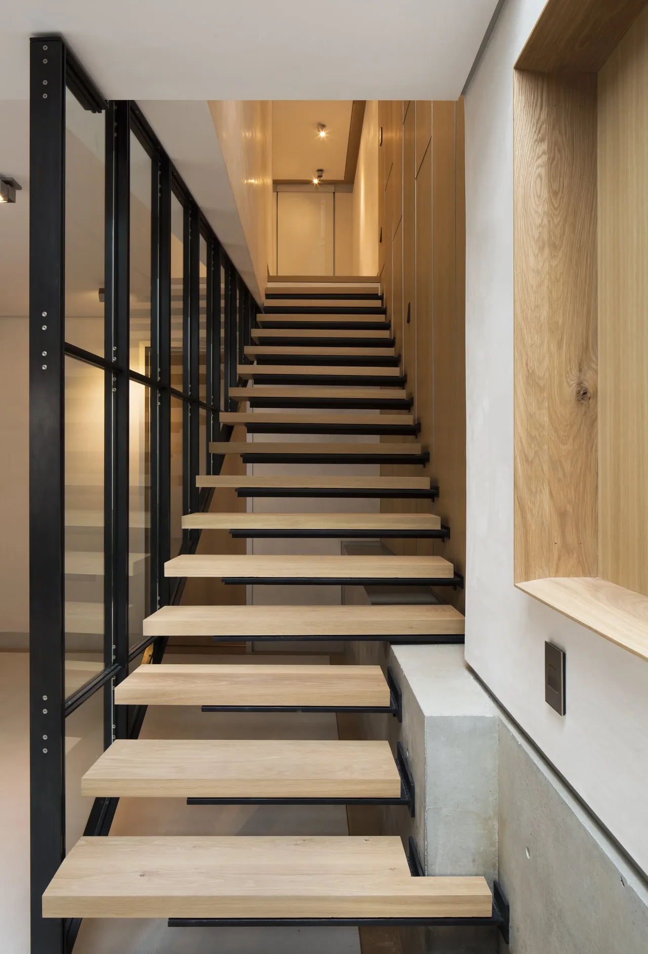 Minimalist floating stair with wood treads and black metal supports beside a wall of black-framed glass panels.