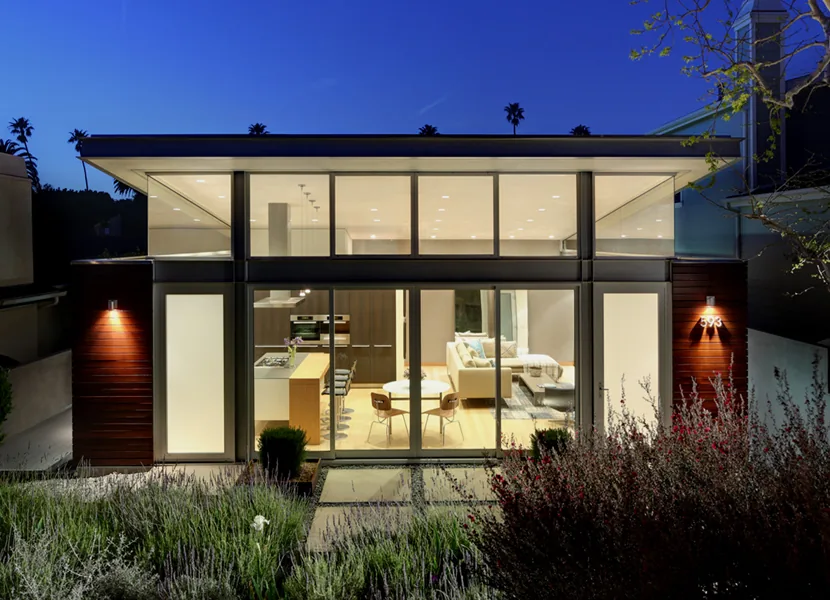 Modern home with extensive glass walls revealing the kitchen and living area, featuring natural wood finishes and front garden.