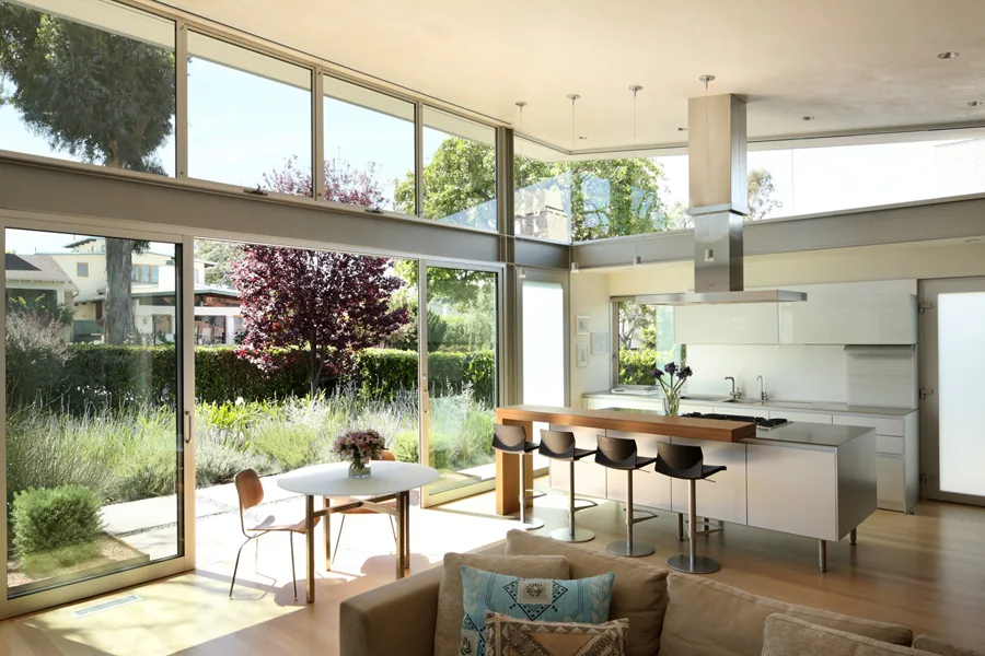 Open kitchen and dining space with glass sliding doors overlooking a lush garden, featuring a kitchen island and bar seating.