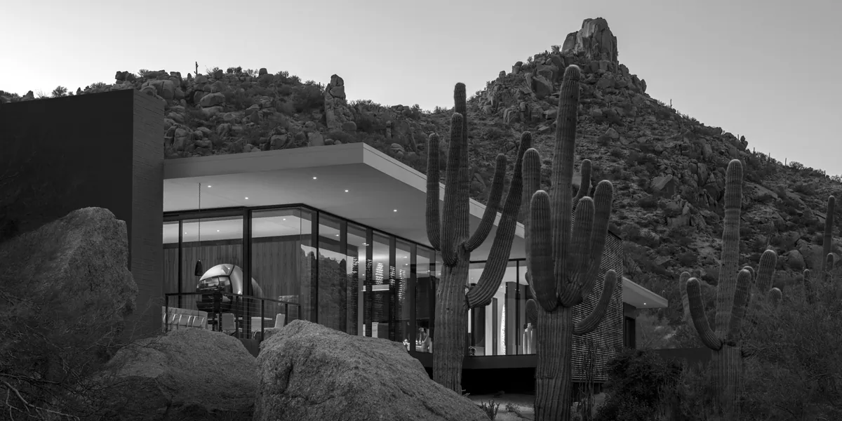 Modern desert home featuring floor-to-ceiling glazing and a flat roof, nestled among desert rocks and tall cacti.