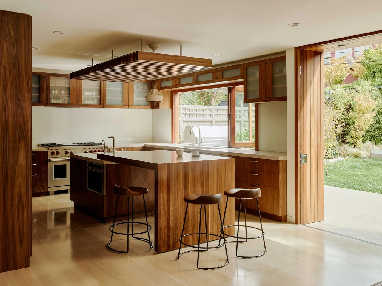 The kitchen features a built-in wooden island with bar stools and opens to a landscaped yard through wide wooden doors.