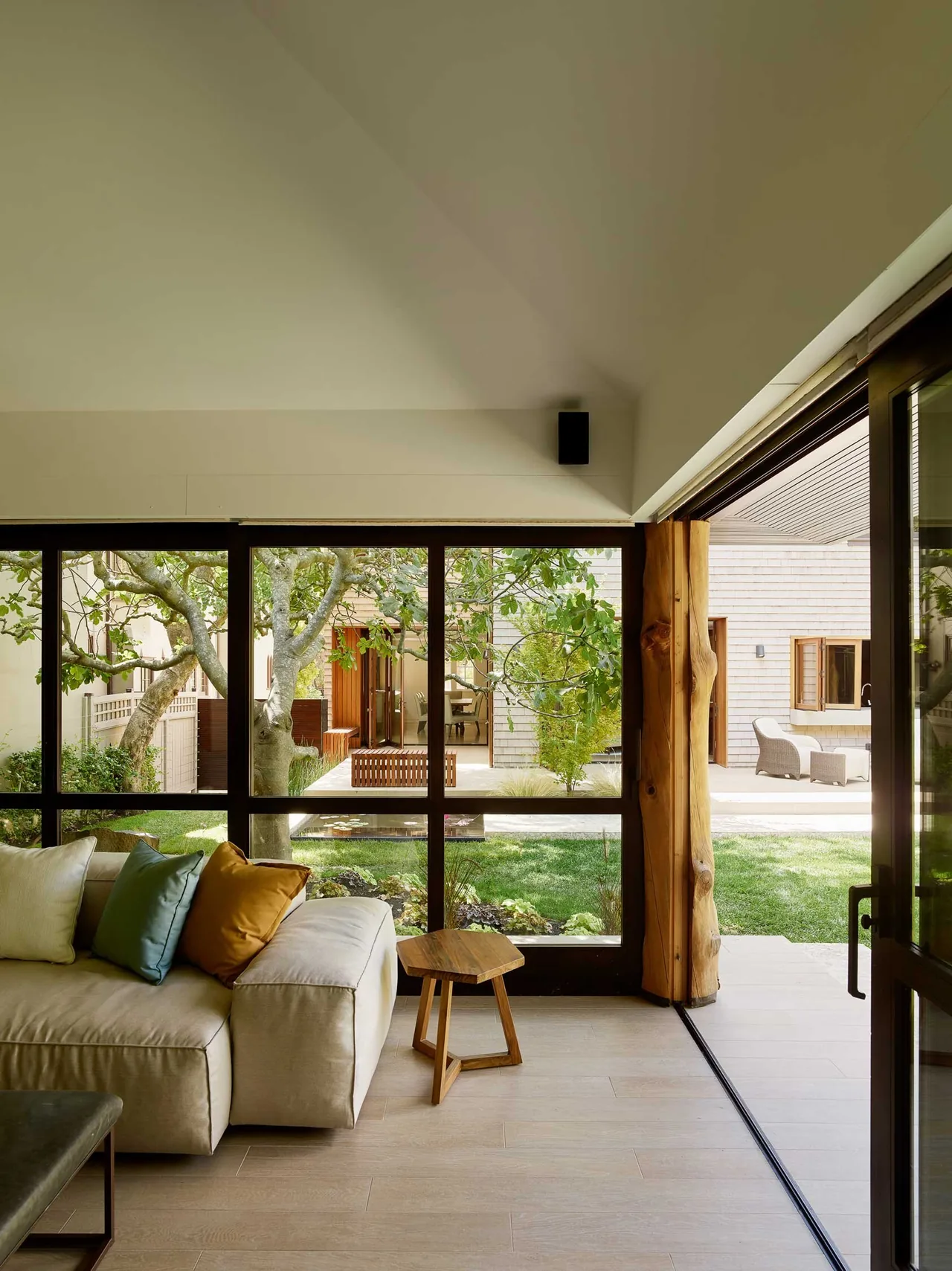 Living room opens through large black-framed windows and sliding door to a green garden and terrace with wood furniture.