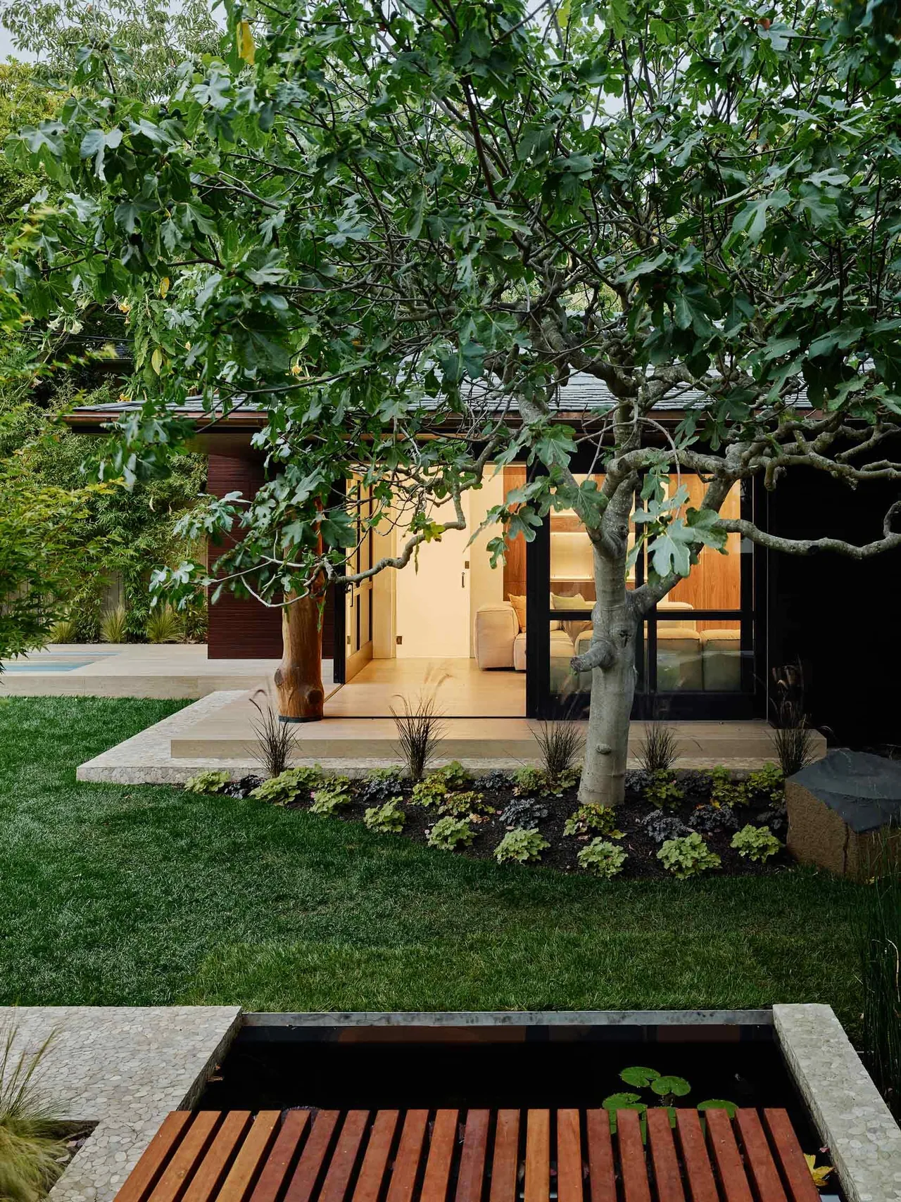 Backyard and garden area of the Claremont Residence with mature tree, pond, wooden bridge, and view into the living space.
