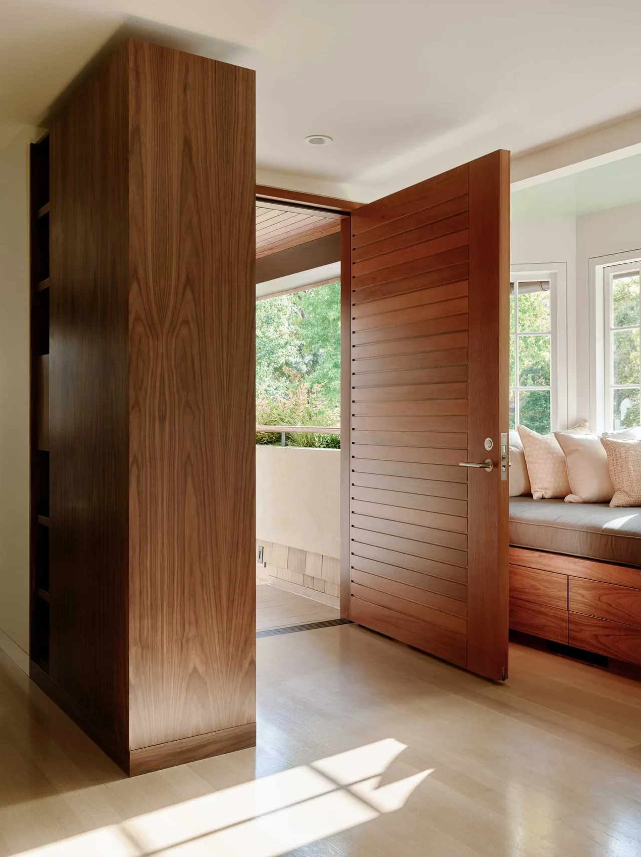 Wooden pivot door opens to a light-filled room featuring built-in bench seating with cushion and wood cabinetry below windows.
