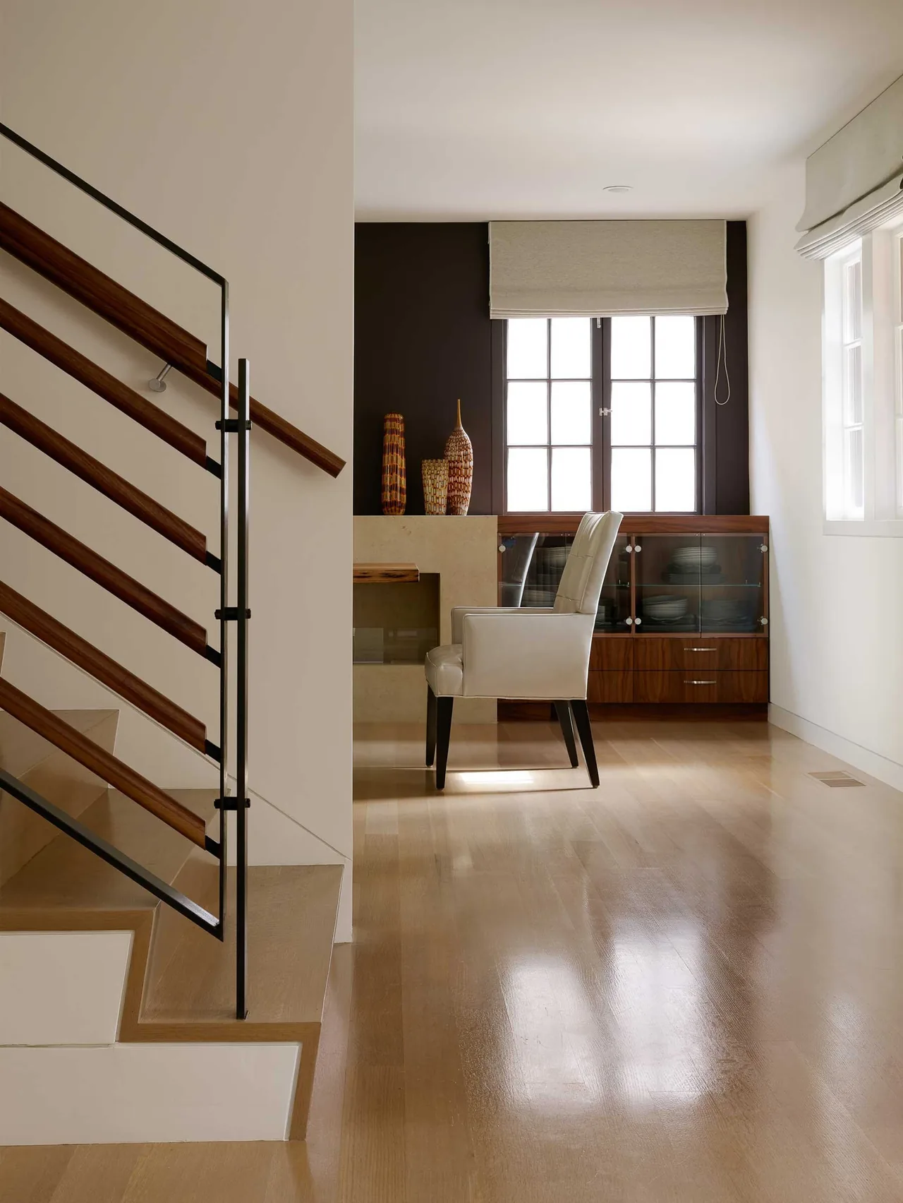 Interior view showing a wooden staircase and a beige armchair near stained wood and glass cabinetry under a window.