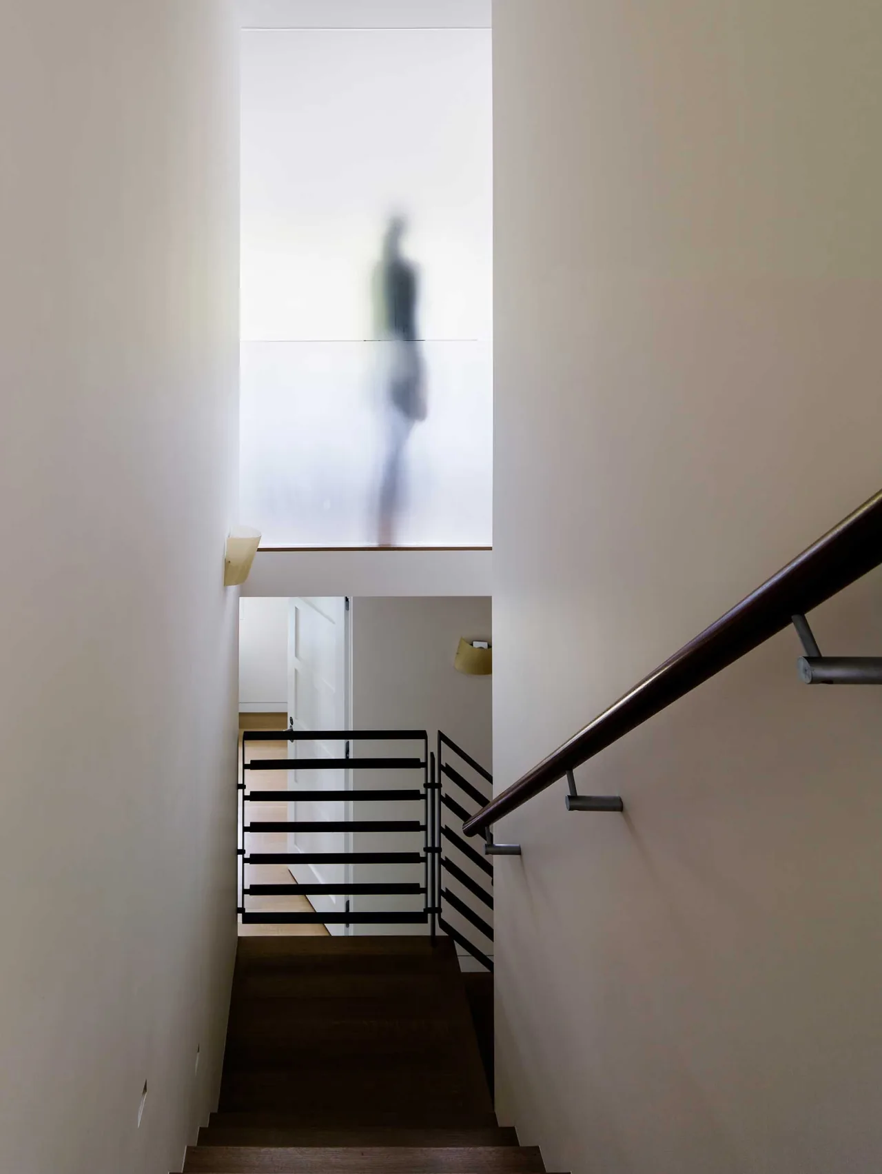 Staircase with wooden handrail and metal railing leading down to a hallway with a frosted glass wall above, featuring a shadowy figure.