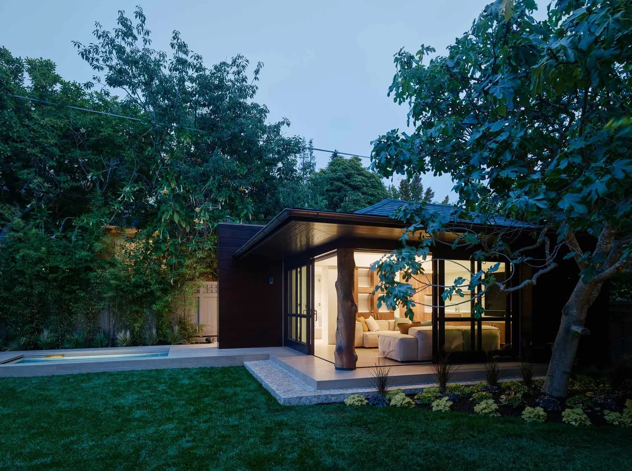 Evening view of a modern single-story home with large glass windows opening to a landscaped yard and a tree in the foreground.