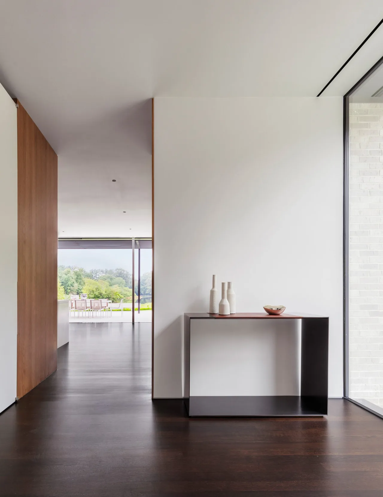Hallway interior shows dark wood flooring, white walls, a minimal metal console table, and a glimpse of outdoor patio seating in New Canaan Pavilion.