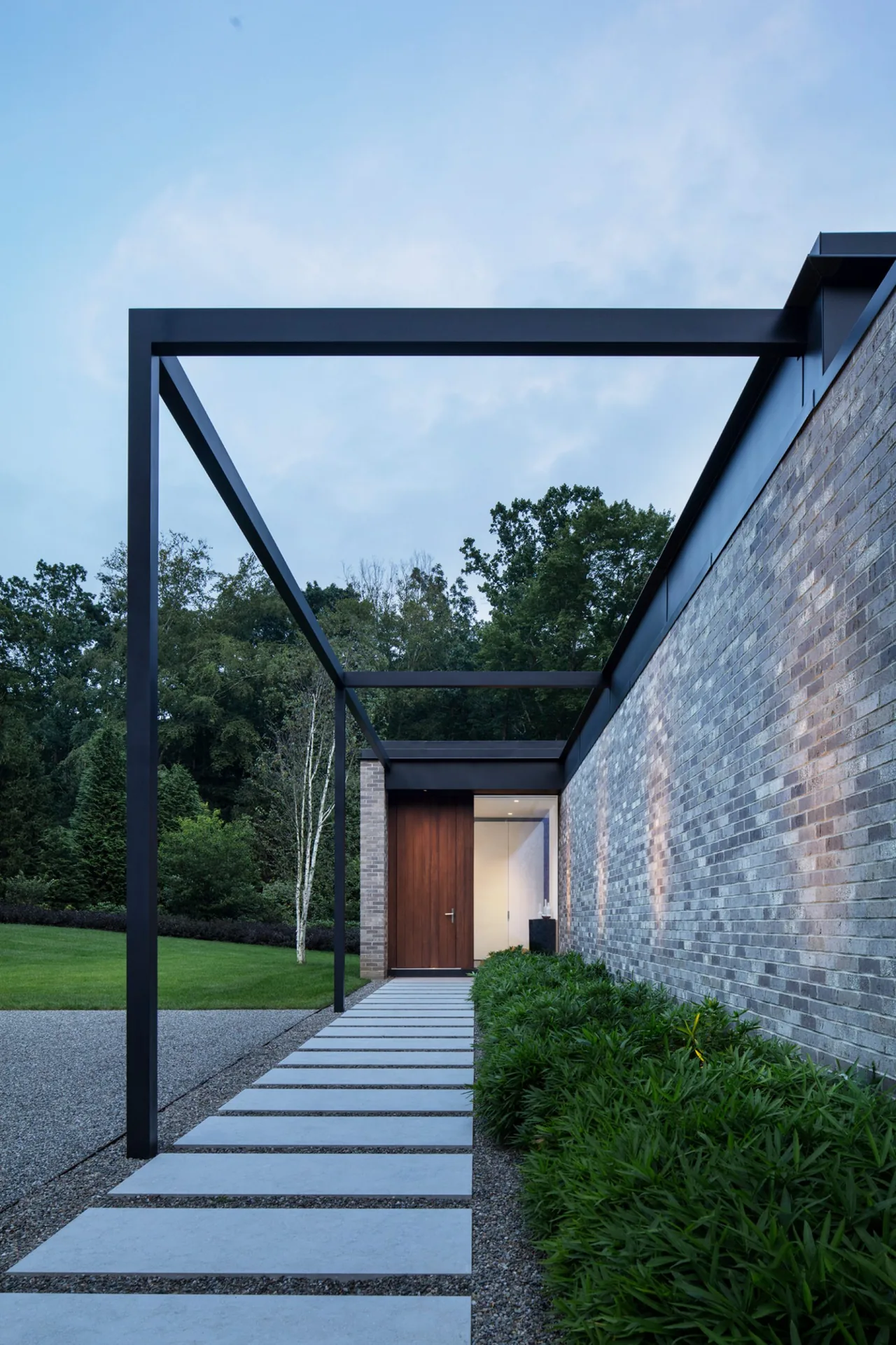 Entry pathway with concrete stepping stones under a black steel pergola structure beside a brick wall in a green lawn setting.