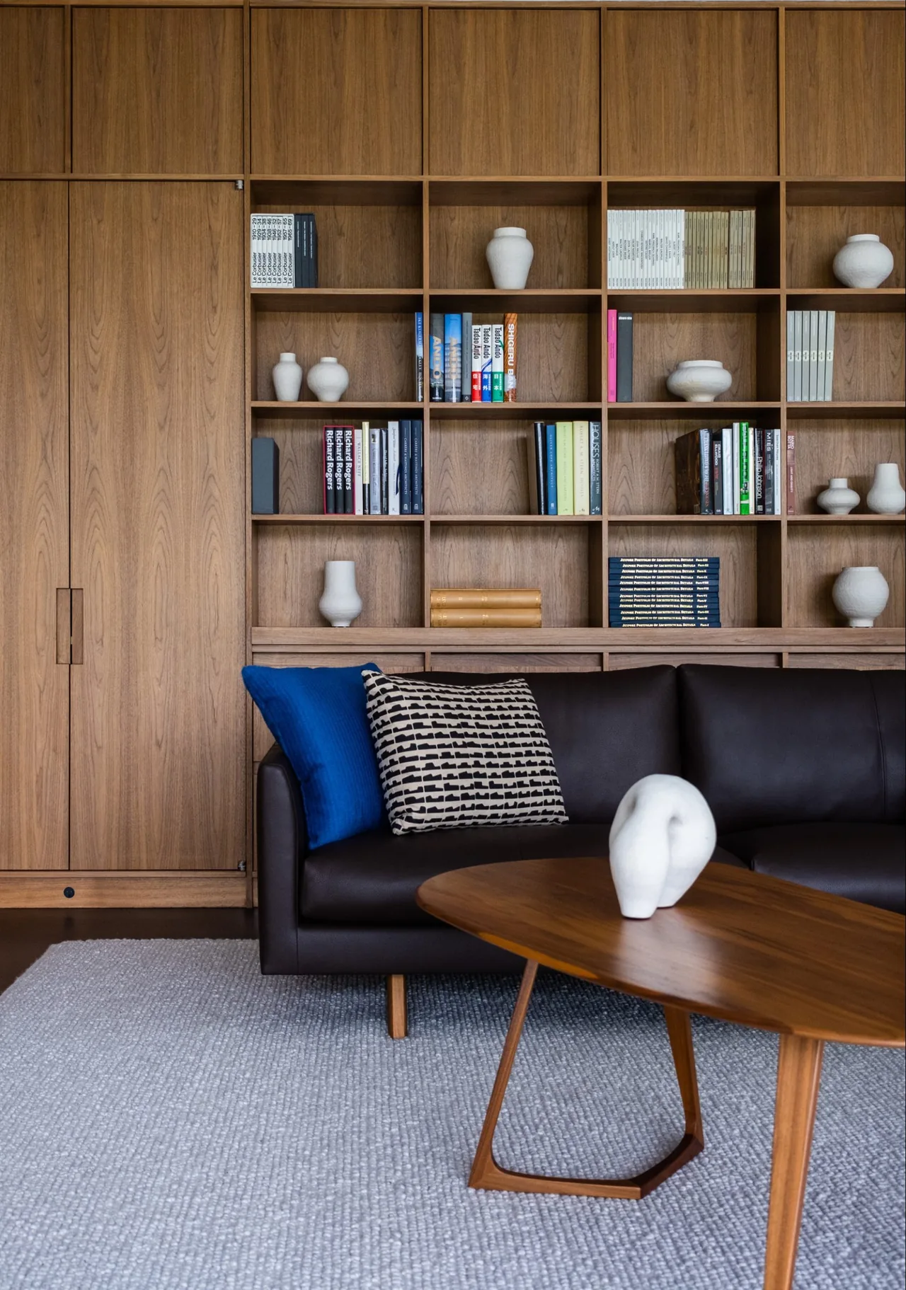 Living room featuring built-in wood shelving, a dark leather sofa with blue and patterned pillows, and a wooden coffee table with a white sculpture.