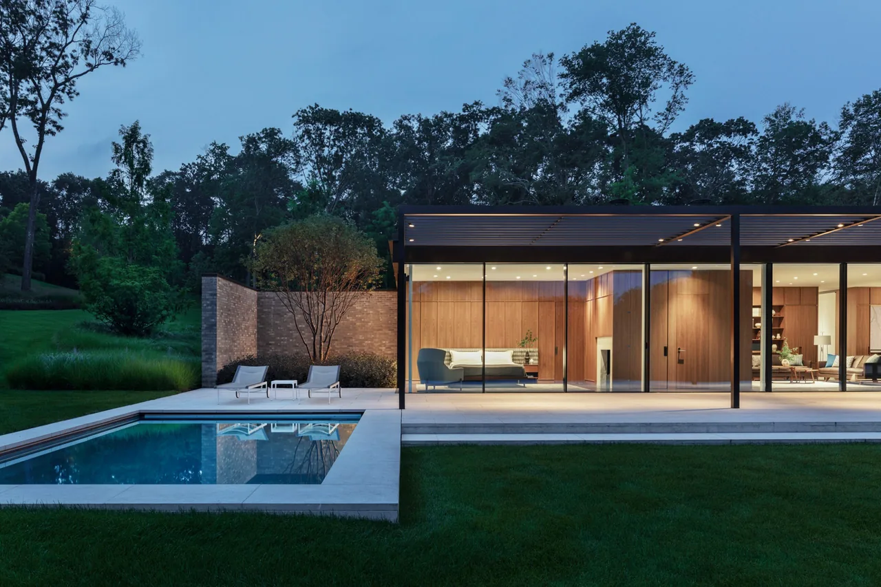 New Canaan Pavilion features floor-to-ceiling glazing and wood paneling, with a pool and outdoor seating area visible at dusk.