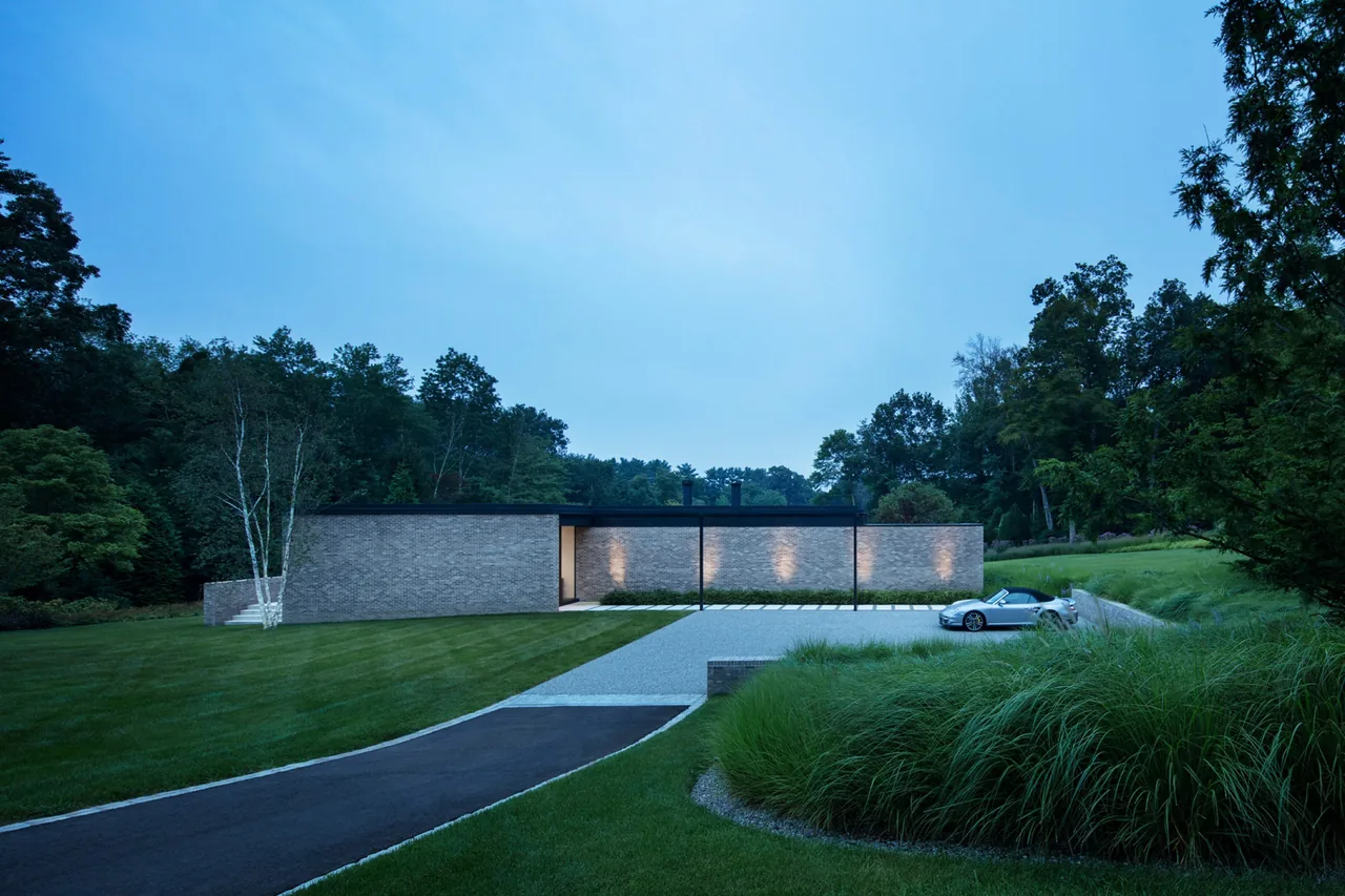 The New Canaan Pavilion is a modern, single-story brick building set in a natural landscape, shown here at dusk with exterior lighting.