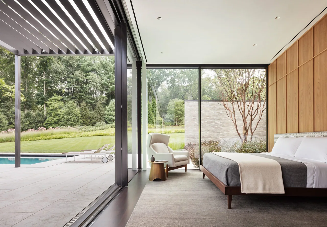 The bedroom features floor-to-ceiling glazing and wood paneling with a view of an outdoor pool and lawn area.
