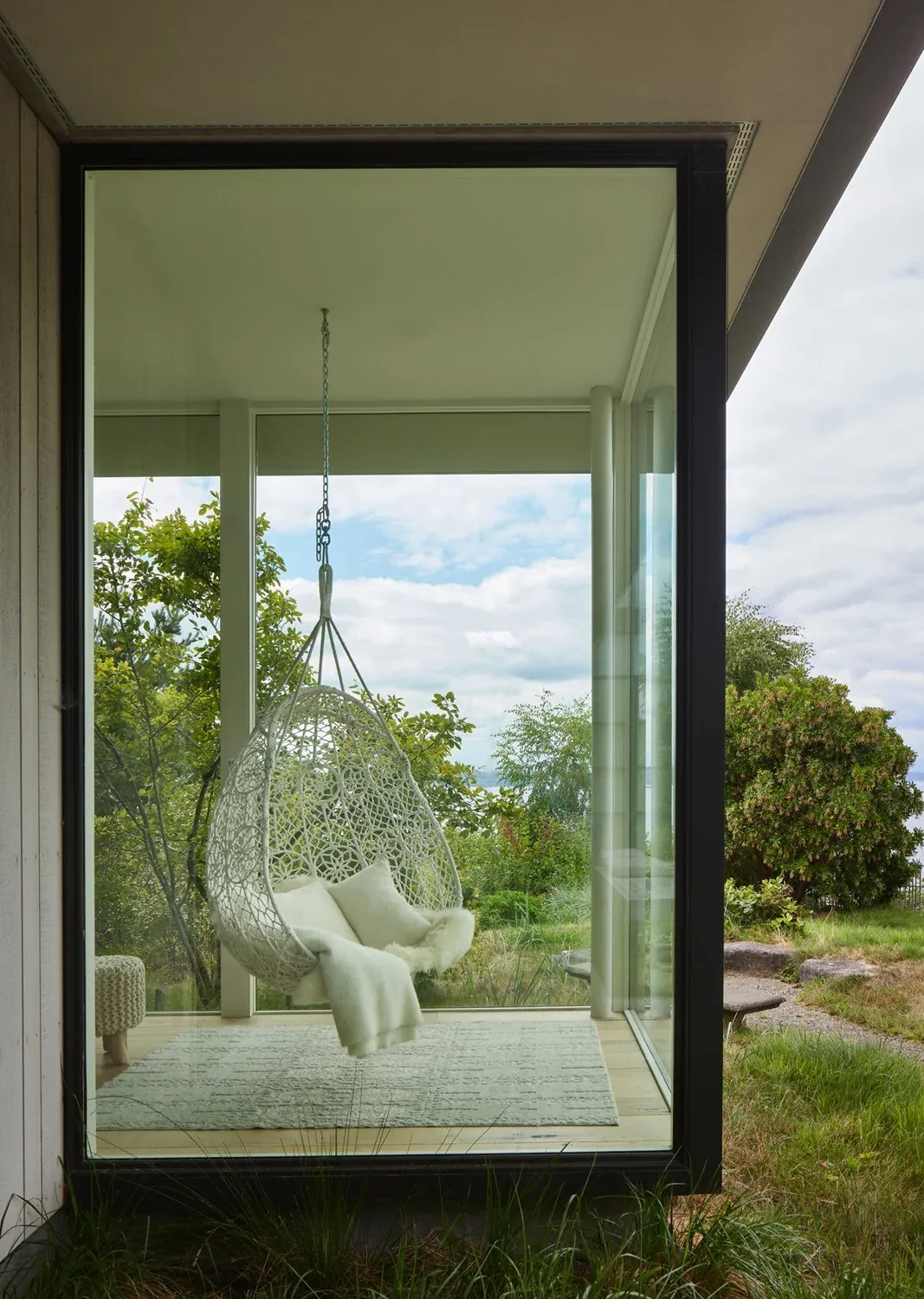 A white woven hanging chair with cushions is suspended inside a glass room with views of the outdoor landscape.