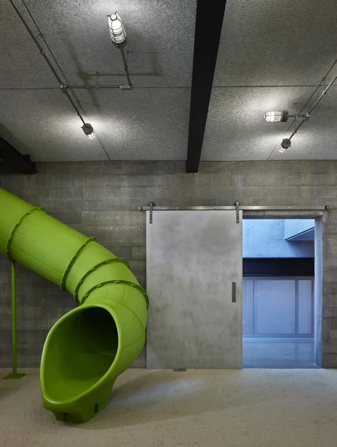 Indoor area features a green spiral slide next to a ribbed concrete wall and a metal sliding door under textured ceiling lighting.