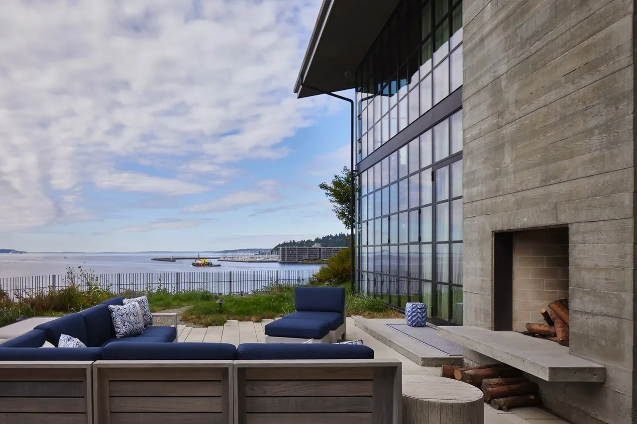 Patio with sectional seating and concrete fireplace beside large glass facade with water view.