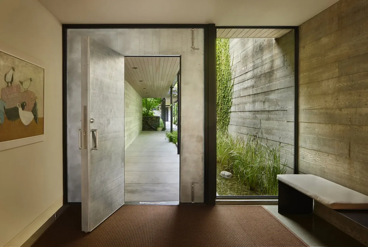 The Bluff features a metal front door with riveted details opening to a concrete exterior corridor and a window with black frame showing a planted courtyard.