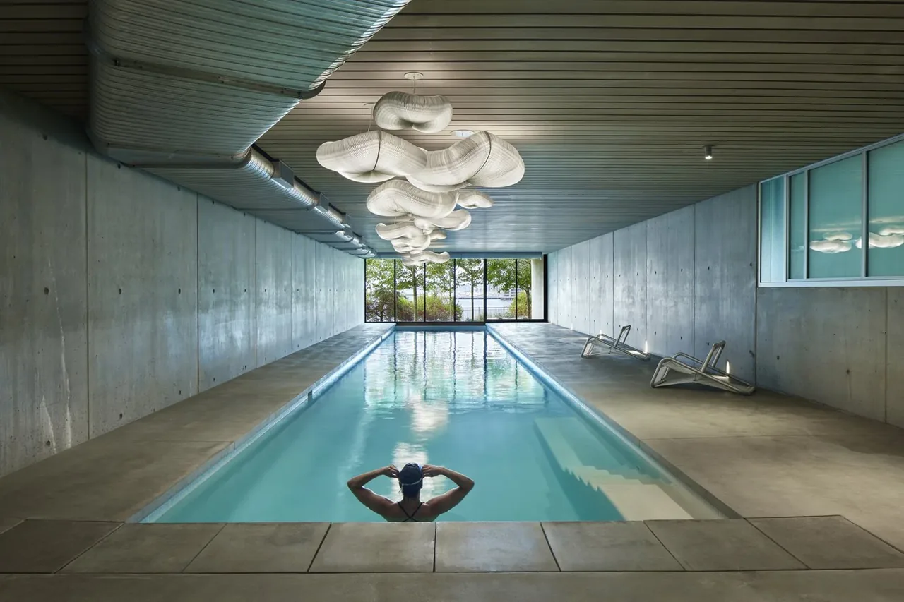 Indoor pool room with exposed concrete walls and ceiling, featuring sculptural pendant lights and reclining pool chairs.