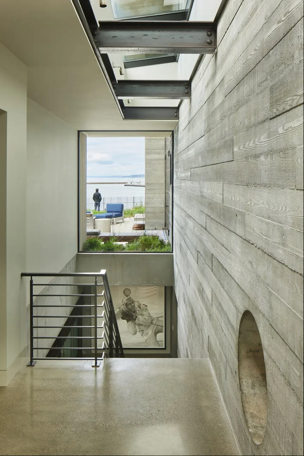 Stairway with metal railing adjacent to a concrete wood-grain textured wall and large window overlooking outdoor seating and waterfront.