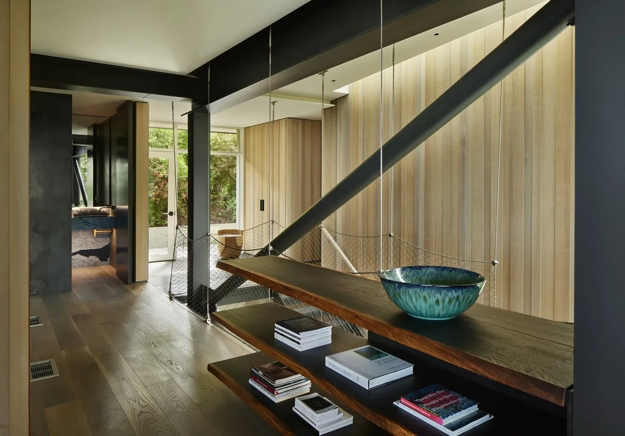 The hallway of Creek House features wood flooring, floating shelves with books, and vertical wood paneling with a metal brace.