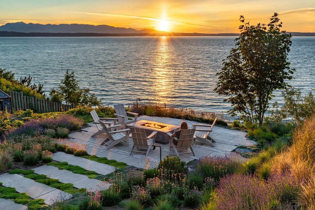 Concrete patio with wooden Adirondack chairs and a fire pit set in landscaped garden by the water at sunset.