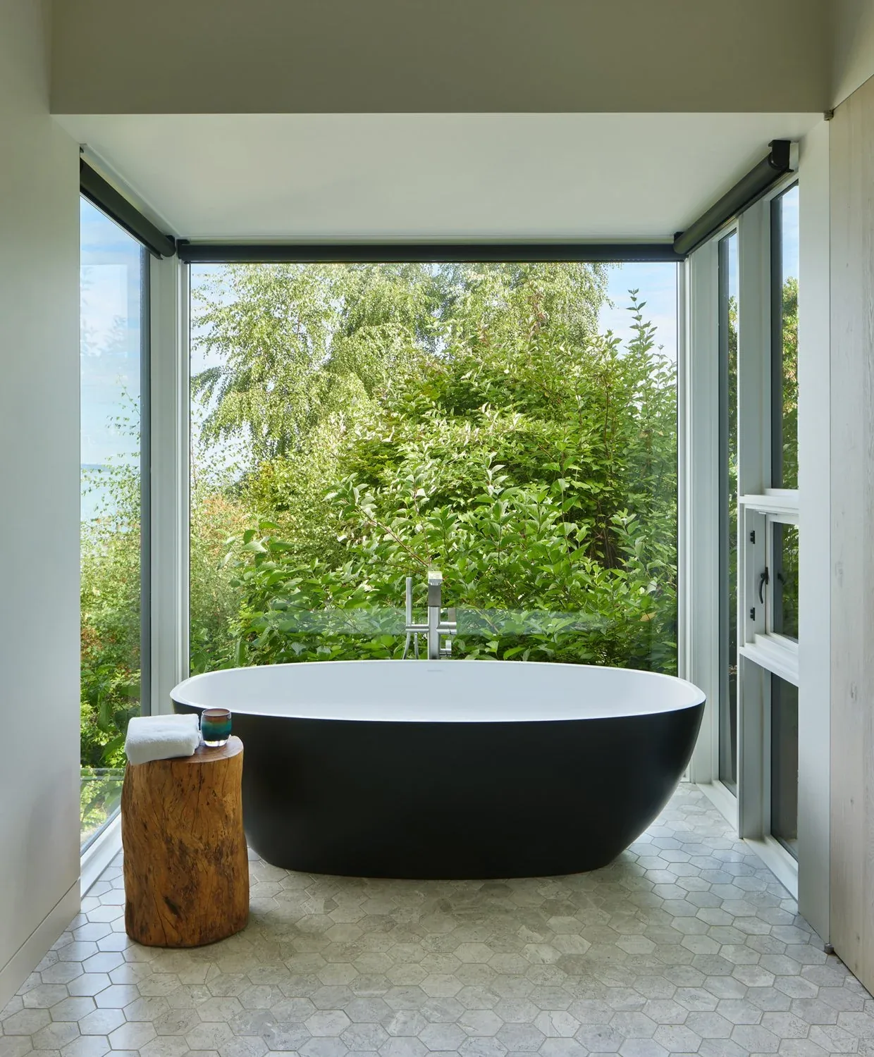 Bathroom with black freestanding tub and large windows overlooking lush greenery at Creek House by DeForest Architects.