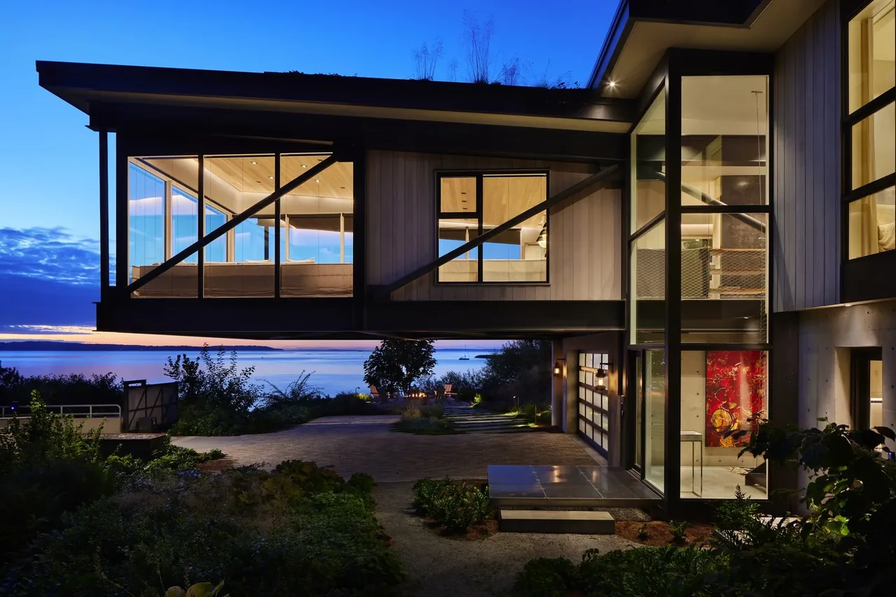 Cantilevered upper floor with black window frames offers expansive views of water at dusk in Creek House.