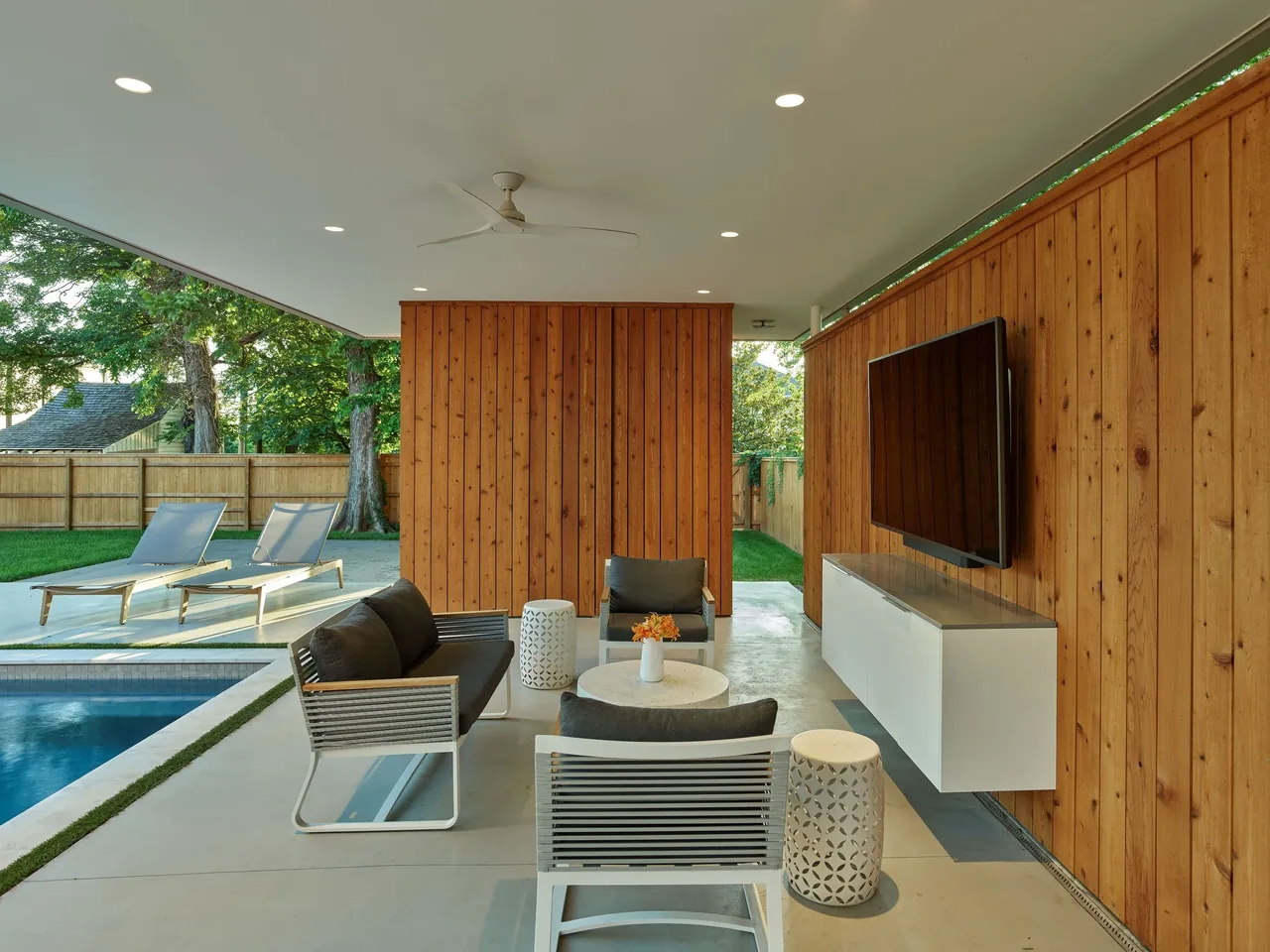 Outdoor pavilion featuring wood siding, a mounted TV, and outdoor seating adjacent to a pool with lounge chairs.