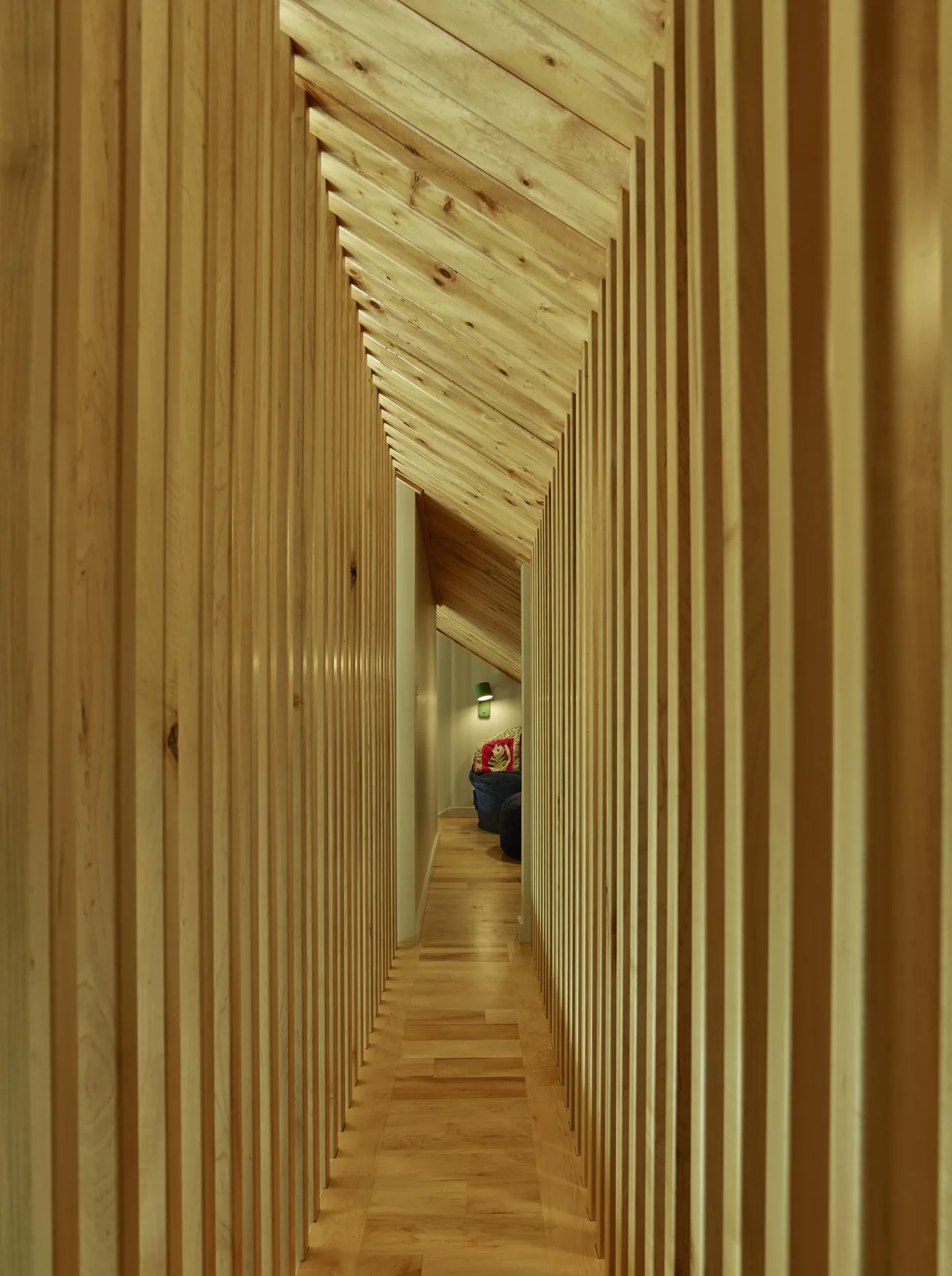 Narrow corridor featuring vertical wood slats and an angled wood ceiling ends at a cozy seating area with a wall sconce.