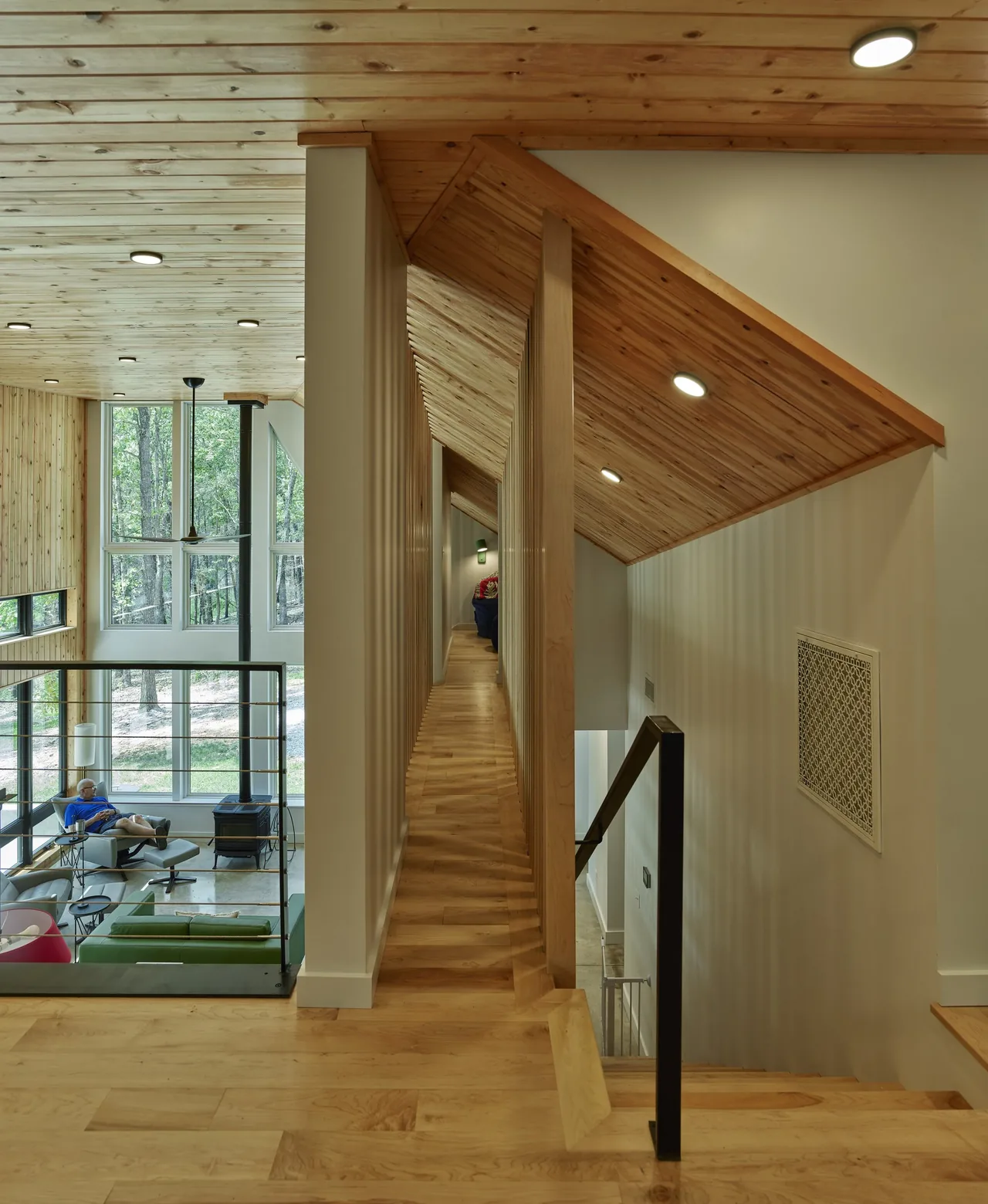 Modern home's upper hallway with wood paneling, recessed ceiling lights, and a view into a living room with large windows.