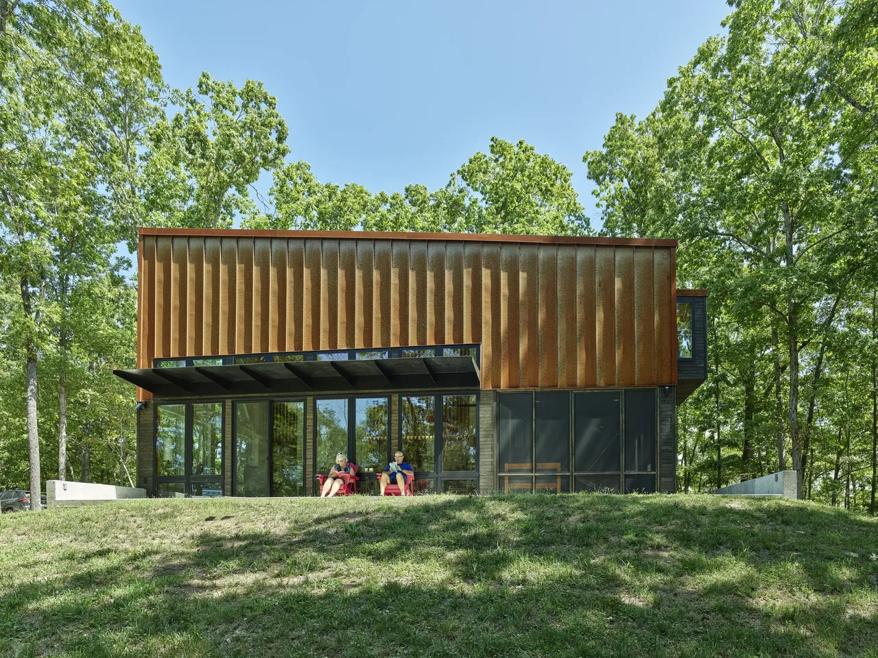 Eclipse Ridge house featuring rust facade and large glass windows with a covered patio and red chairs.