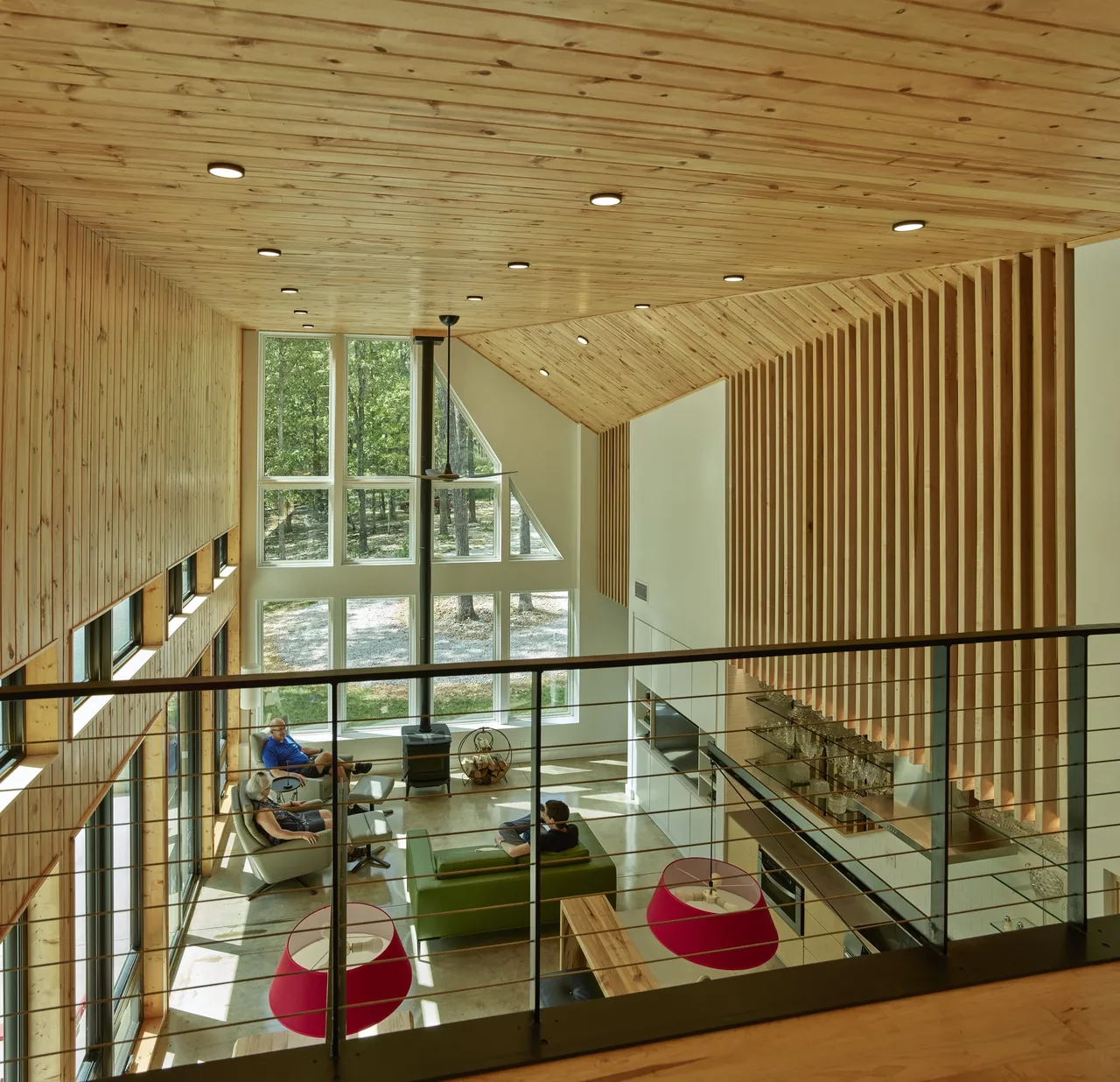 Living room with pine wood slat ceiling and large picture windows, featuring a green sofa and wood-burning stove.