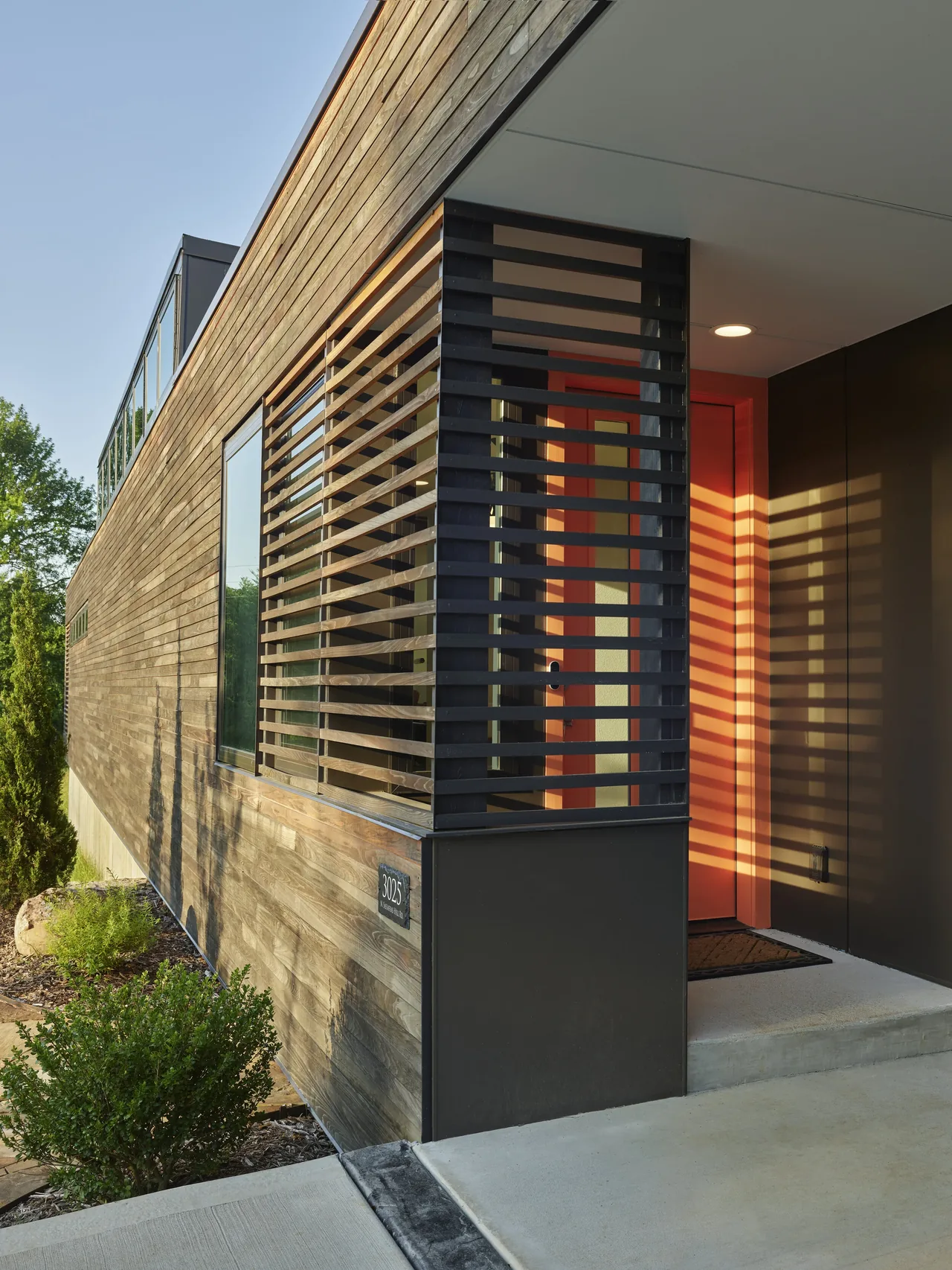 The Willis Residence features wood siding with a black metal slatted privacy screen near its red front door entry.