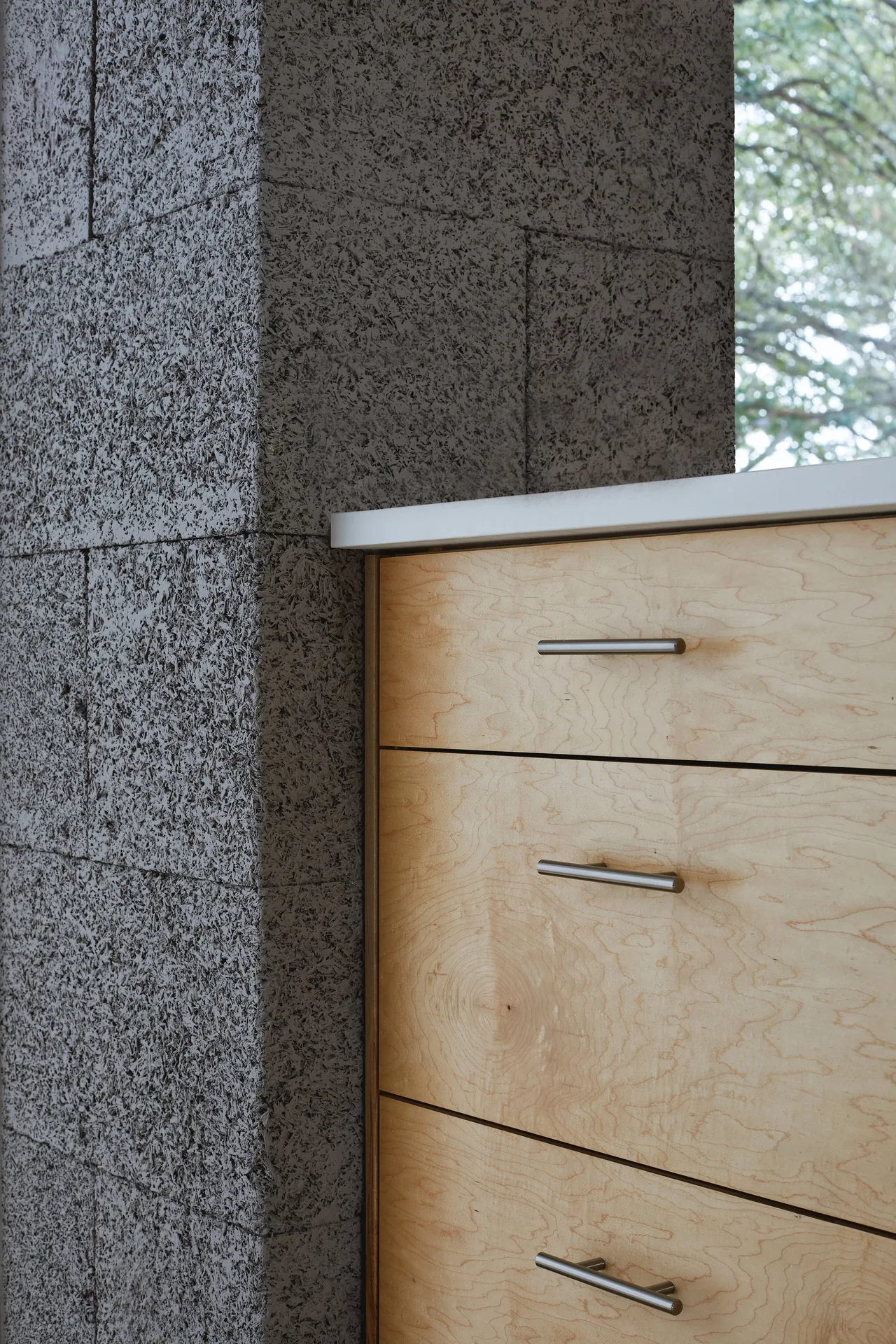 Light wood kitchen drawers sit alongside a textured concrete block wall next to a window with tree views.