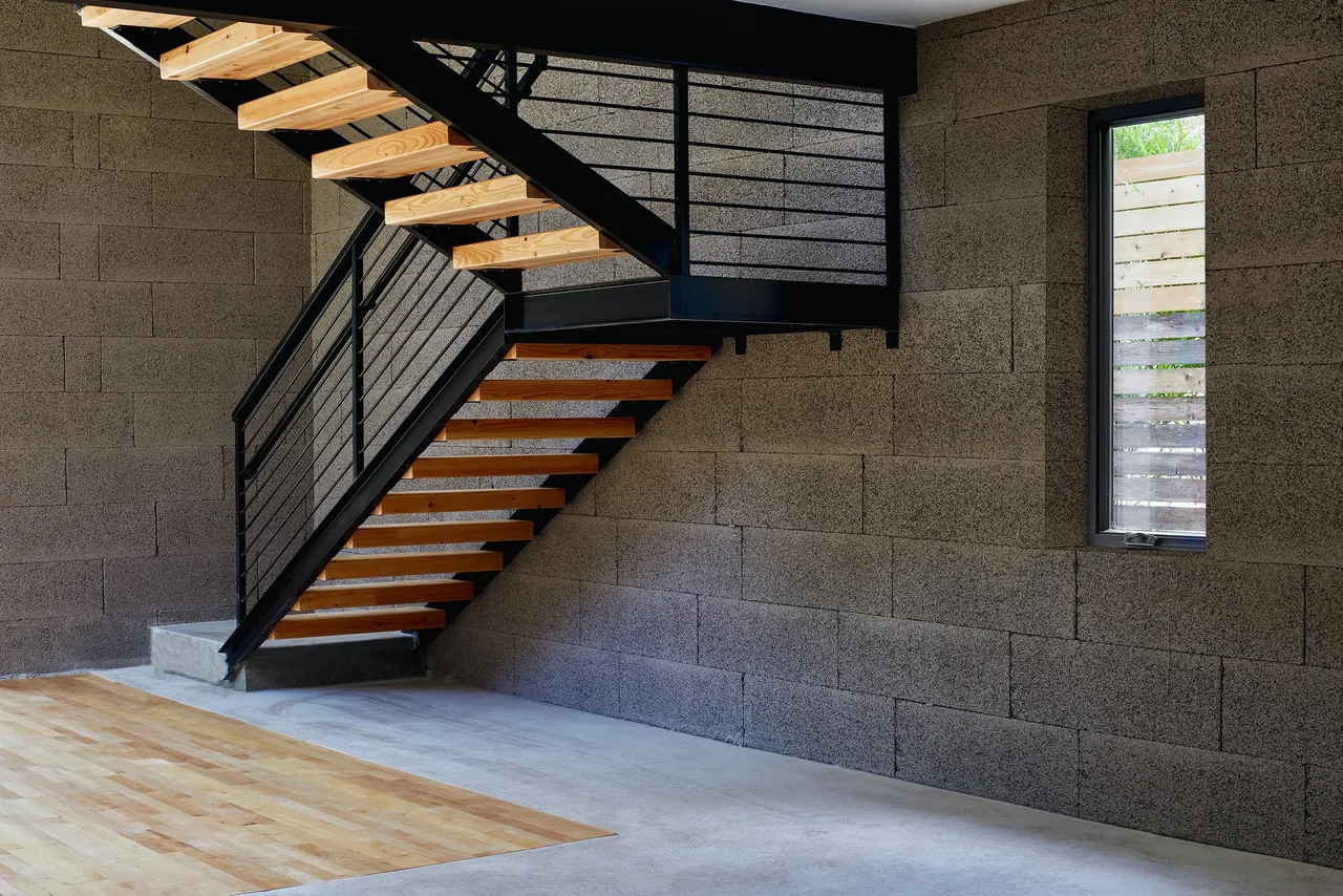 Open riser staircase with wood treads and black railings adjacent to textured concrete block wall and narrow window.