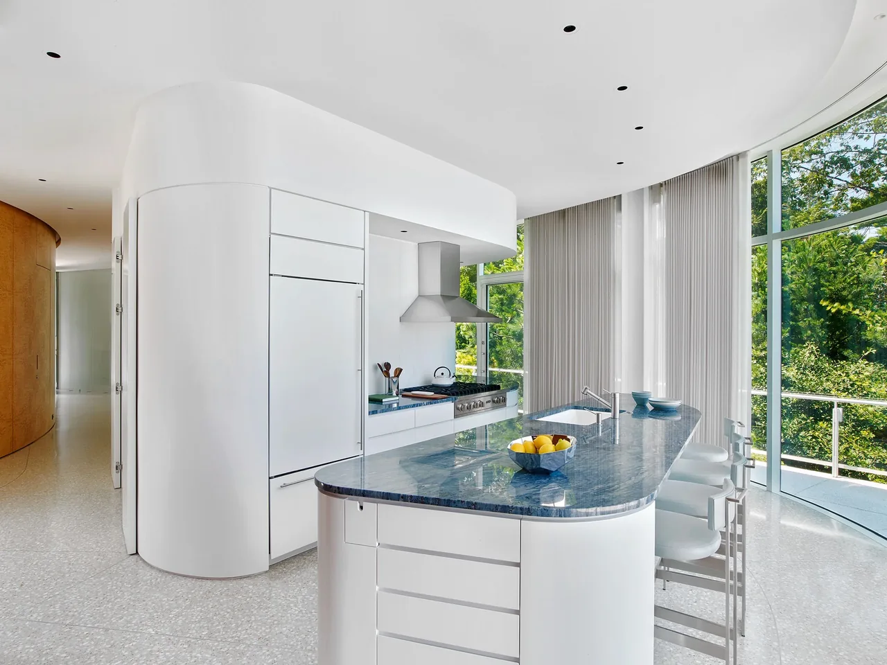 The kitchen features a curved blue stone island with white cabinetry and a curved glass window wall.