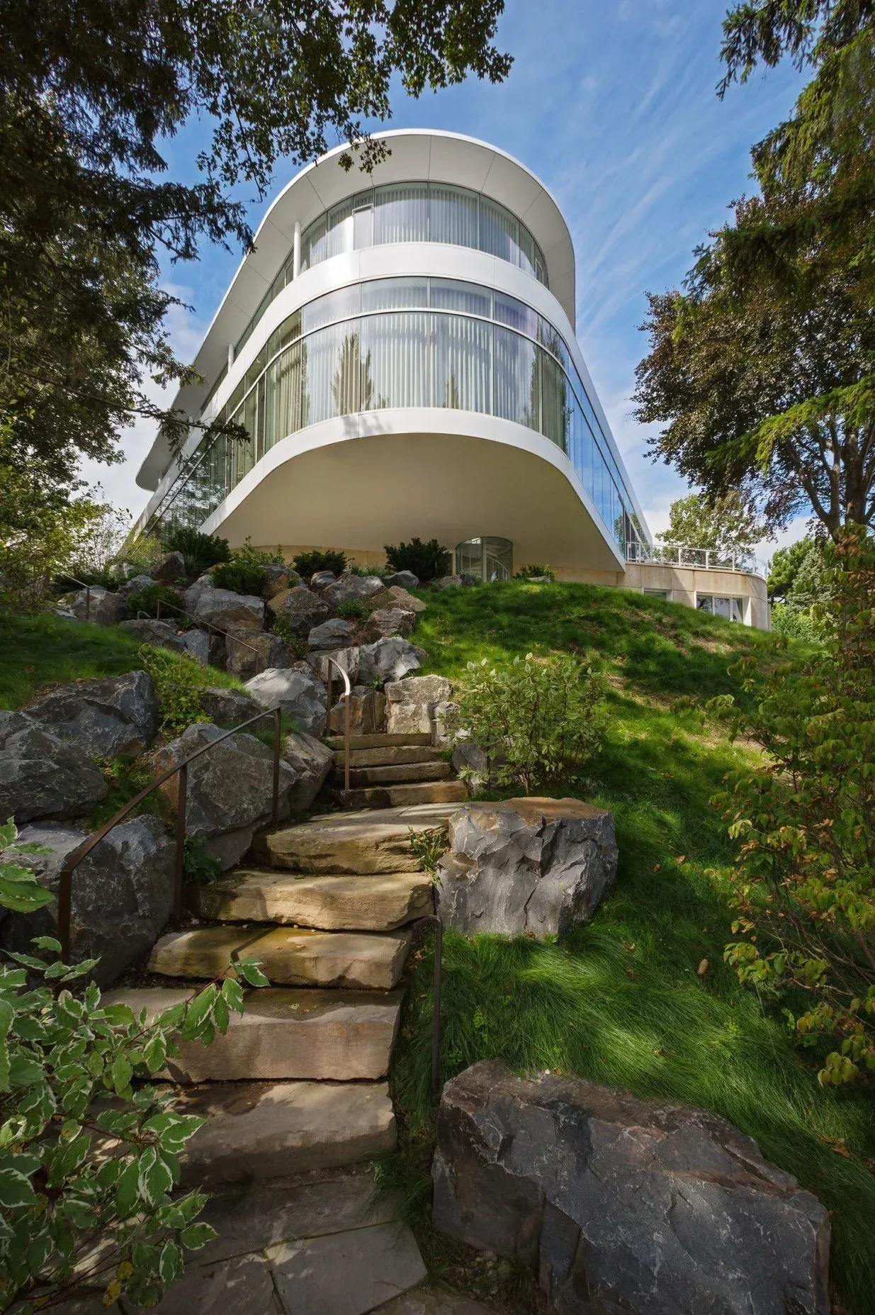 The Lake Michigan Residence features a curved white exterior with floor-to-ceiling glass windows and stone steps leading up a landscaped hill.