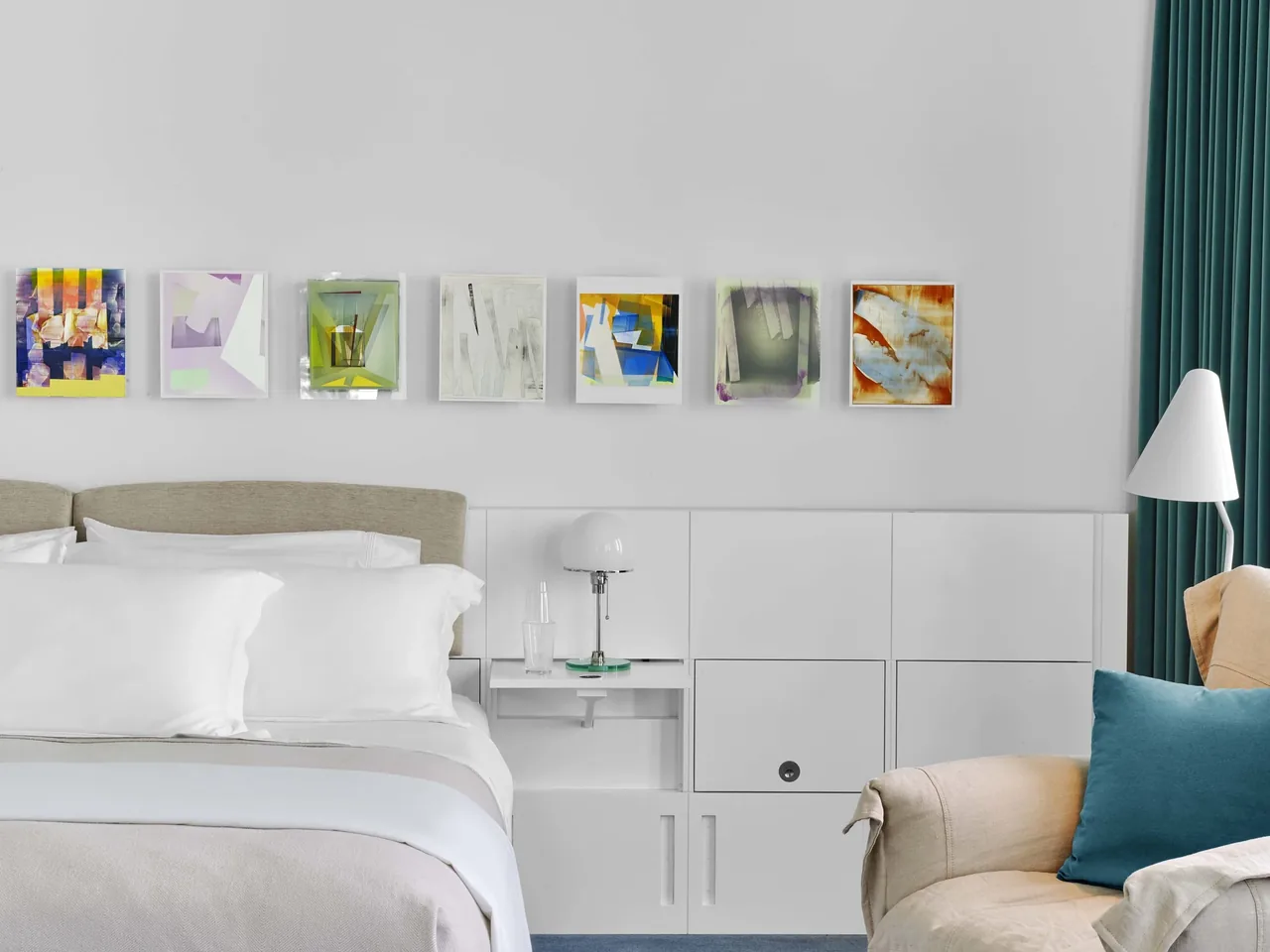 Bedroom in Lake Michigan Residence features white bedding, abstract wall art, and a beige armchair accented by teal curtains and pillow.