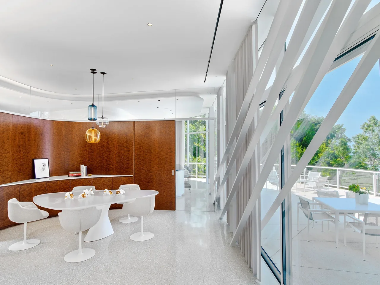 Dining room with a white oval table, curved wood panel wall, pendant lights, and floor-to-ceiling slanted windows with outdoor seating visible.