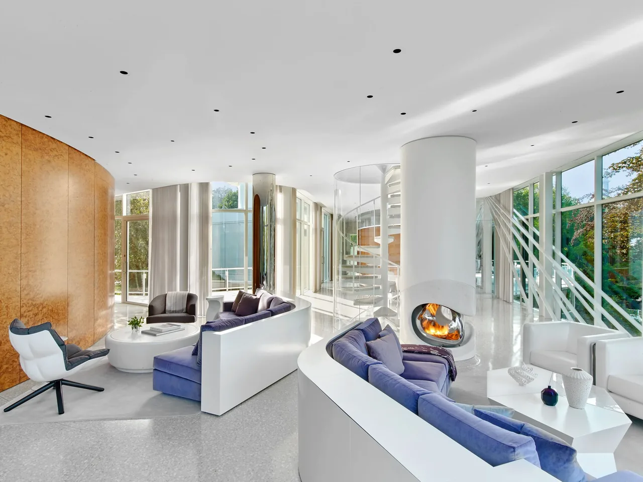 Living room features curved white sofas, a central cylindrical fireplace, and extensive floor-to-ceiling glazing with views of surrounding trees.