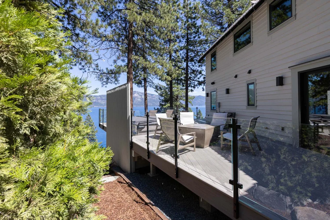 Lake view deck with glass railing and seating area adjacent to a two-story home with white siding.